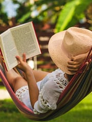 Junge Frau mit Strohhut und Buch liegt entspannt in einer Haengematte im Garten und liest