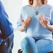 birgit-nora-schaefer-coaching-gespraech-1000x1000 Bildausschnitt aus einer Coaching-Szene, in dem sich zwei Menschen gegenüber sitzen und eine der beiden Frauen im blauen Pullover mit einer silbernem Kette und großem ziselierten Silberanhänger die Hände in einer sprechenden Geste auf sich richtet