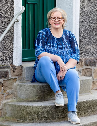 birgit-nora-schaefer-sitzt-entspannt-auf-einer-treppe-1000x1300 Birgit Nora Schäfer sitzt mit angezogenem rechten Knie lächelnd auf einer Steintreppe auf der Margarethenhöhe in Essen, NRW