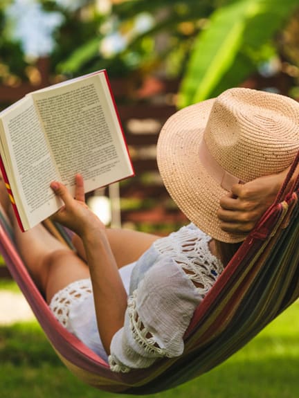 Junge Frau mit Strohhut und Buch liegt entspannt in einer Haengematte im Garten und liest