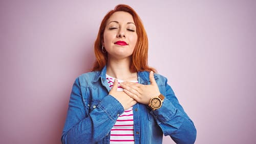 birgit-nora-schaefer-dankbarkeit-empfinden-2000x1125 Junge rothaarige Frau mit rotem Lippenstift, in rotem Ringelshirt und Jeansjacke legt mit geschlossenen Augen beide Haende auf ihr Herz
