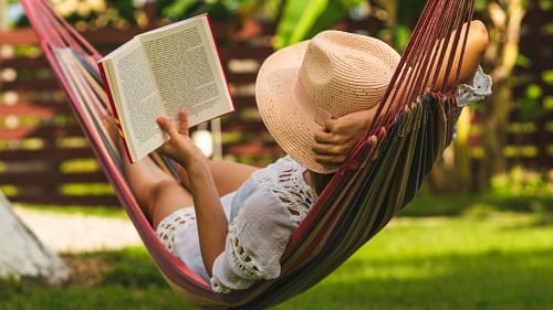 birgit-nora-schaefer-entspannung-in-der-haengematte-2000x1125 Junge Frau mit Strohhut und Buch liegt entspannt in einer Haengematte im Garten und liest