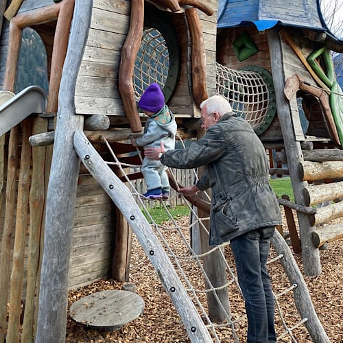 598D9CBC-16AB-4758-84EA-ACD688F7486E Großvater hilft Kind auf Holzspielplatz klettern